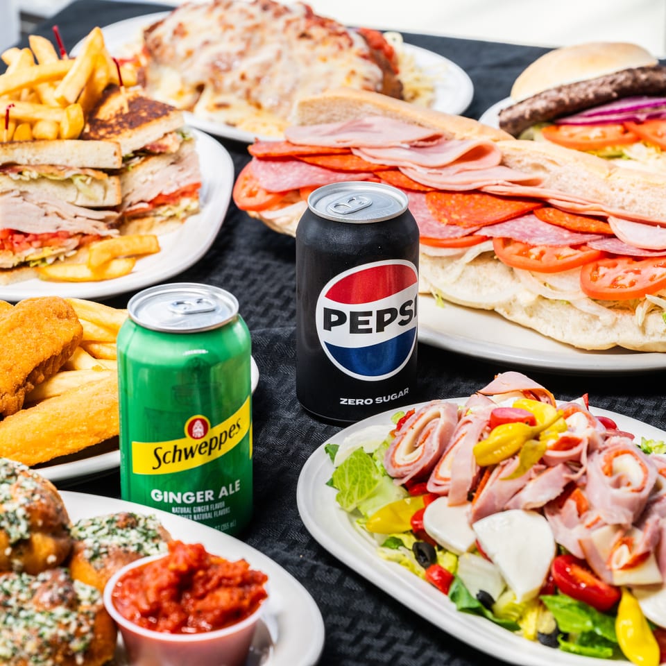 Full table of restaurant food: sandwiches, burger, fried fish, antipasto salad, garlic knots, and sodas.