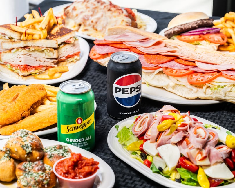 Full table of restaurant food: sandwiches, burger, fried fish, antipasto salad, garlic knots, and sodas.