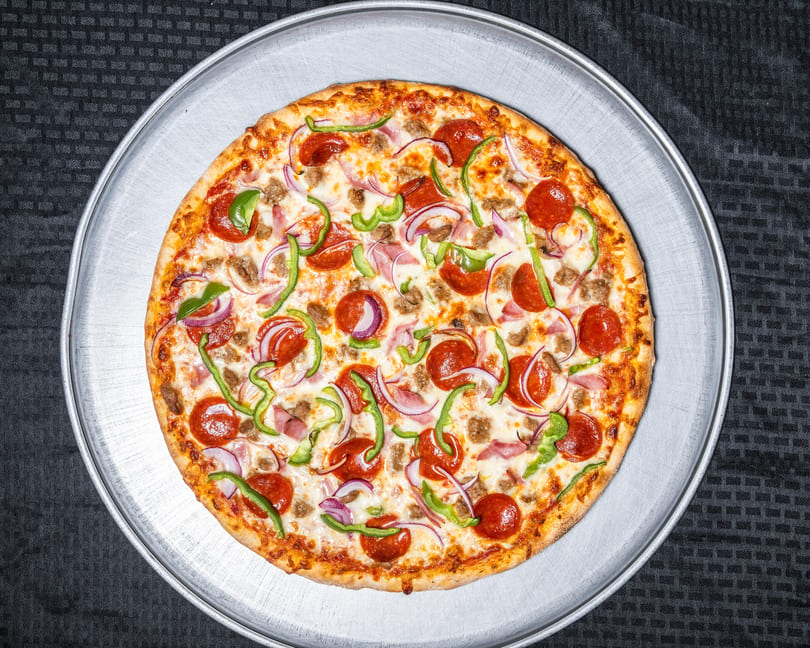 A whole pizza with pepperoni, sausage, ham, green peppers, and red onions on a metal pan.