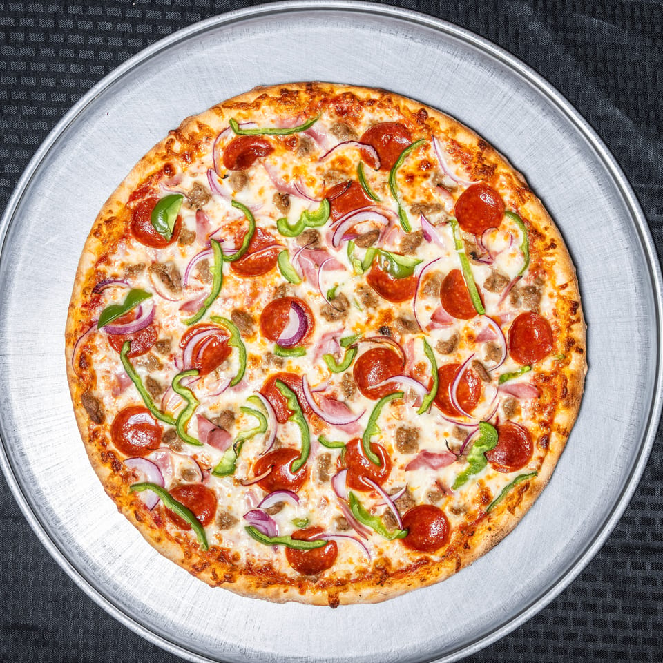 Whole pizza with pepperoni, green peppers, red onions, sausage, and ham on a metal tray.