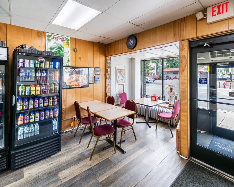 Casual restaurant interior with wood-paneled walls, tables, maroon chairs, a drink cooler, and a street view.