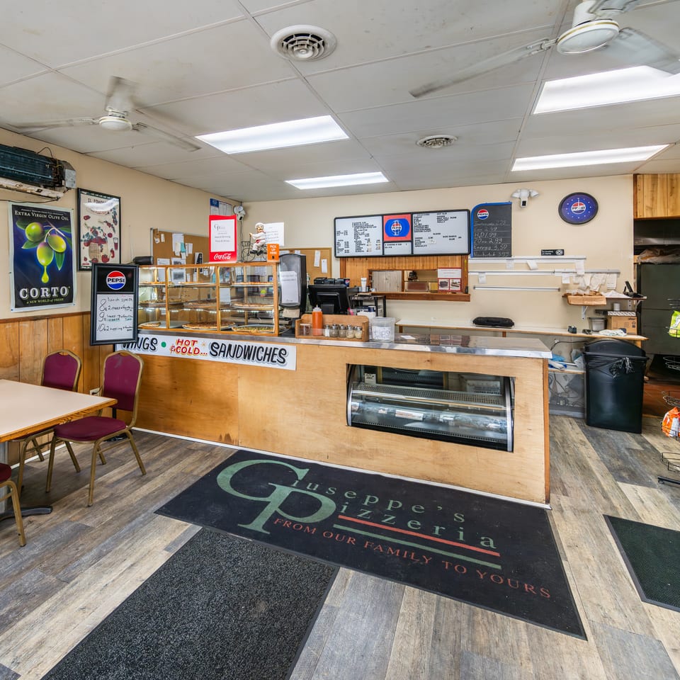 Casual pizzeria interior with a wooden counter, pizza display, menu board, and a small table with red chairs.