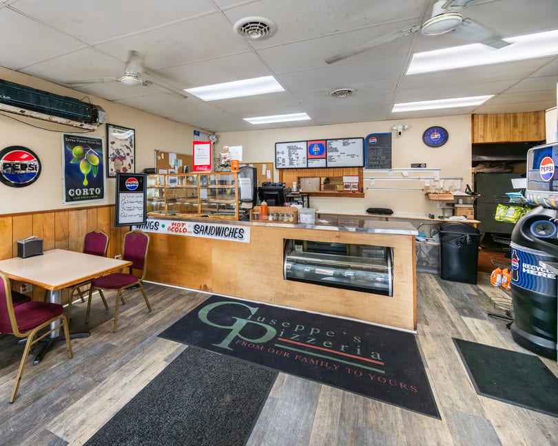 Casual pizzeria interior with a wooden counter, pizza display, menu board, and a small table with red chairs.
