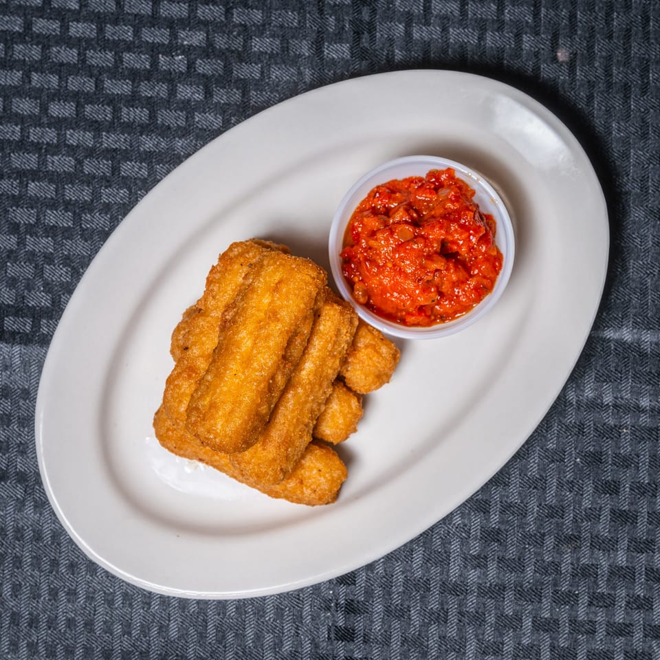A white oval plate holds golden fried sticks and a small cup of red pepper sauce.