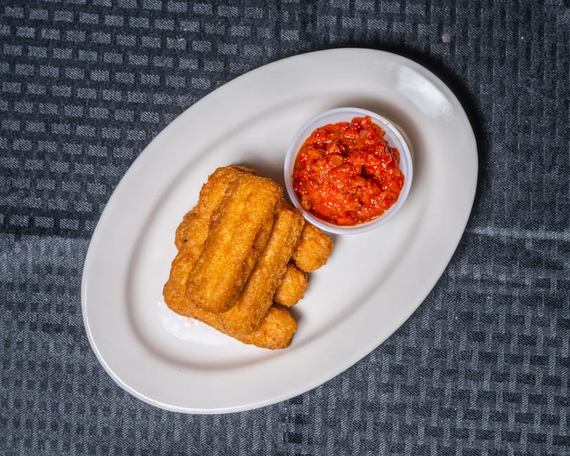 A white oval plate holds golden fried sticks and a small cup of red pepper sauce.
