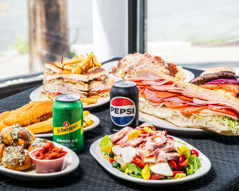 Restaurant table showcasing a variety of dishes: sandwiches, pasta, fried fish, salad, garlic knots, and sodas.
