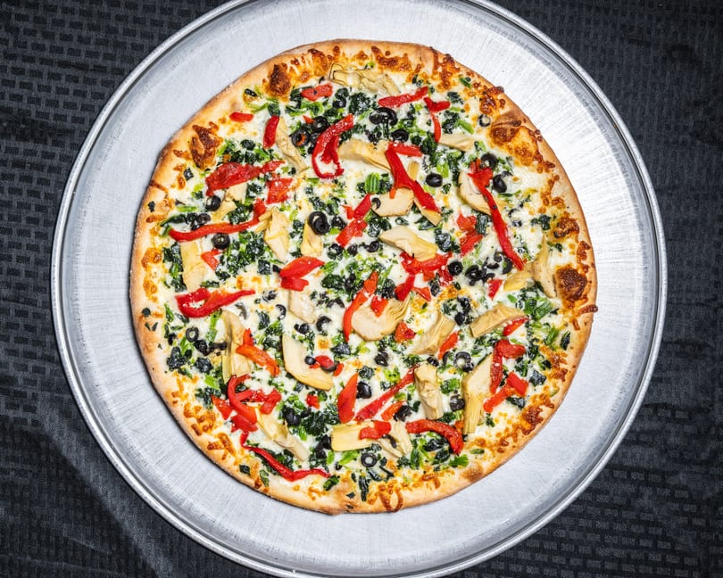 Whole pizza with spinach, black olives, red peppers, and artichoke hearts on a metal pan.