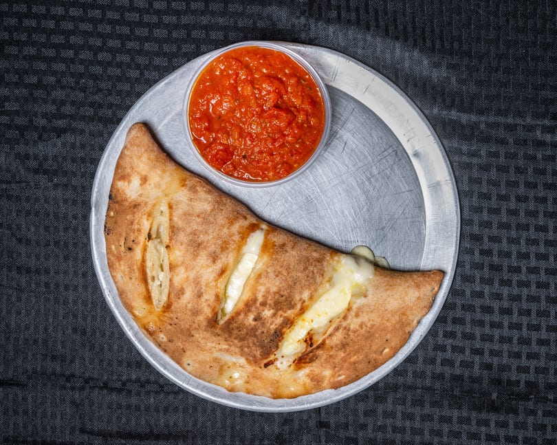 Golden-brown calzone with cheese filling and a side of red marinara sauce on a metal tray.