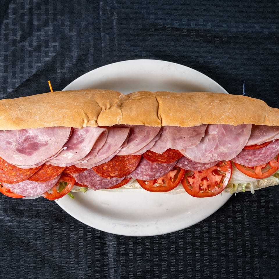 Large sub sandwich filled with ham, salami, pepperoni, sliced tomatoes, and lettuce on a white plate.