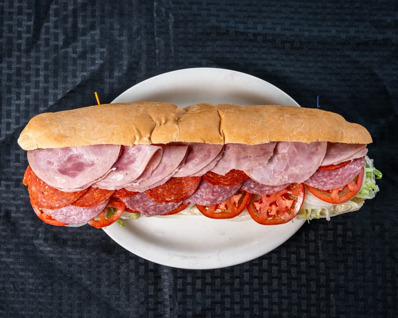 Large sub sandwich filled with ham, salami, pepperoni, sliced tomatoes, and lettuce on a white plate.