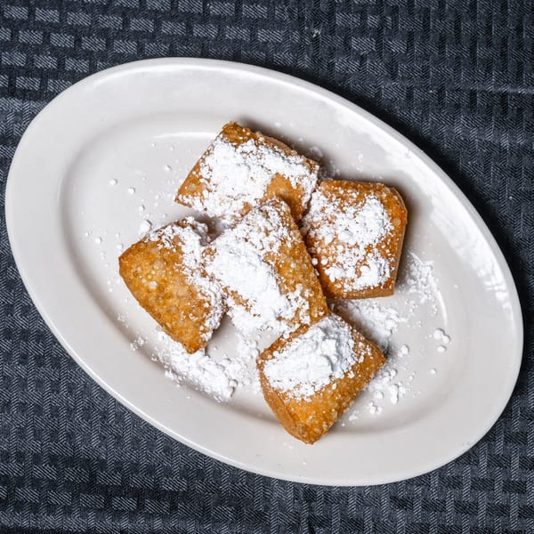 Five golden-brown beignets covered in powdered sugar on a white oval plate.
