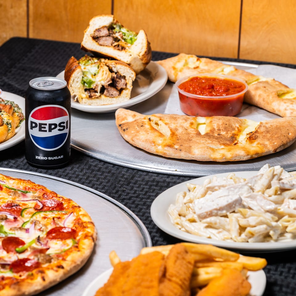 Table filled with pizza, garlic knots, sub, calzones, pasta, chicken tenders, fries, and sodas.