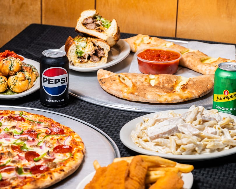 Table filled with pizza, garlic knots, sub, calzones, pasta, chicken tenders, fries, and sodas.