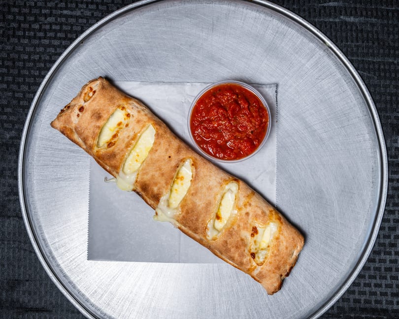 A long, cheesy baked calzone with marinara dipping sauce on a silver pizza pan.