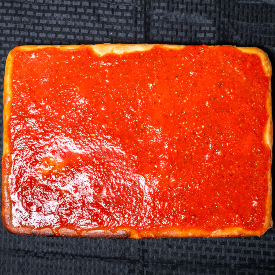 Rectangular pizza with a thick, vibrant red tomato sauce on a dark textured background.