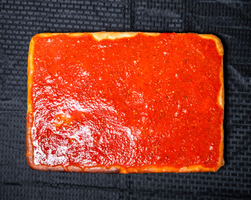 Rectangular pizza with a thick, vibrant red tomato sauce on a dark textured background.