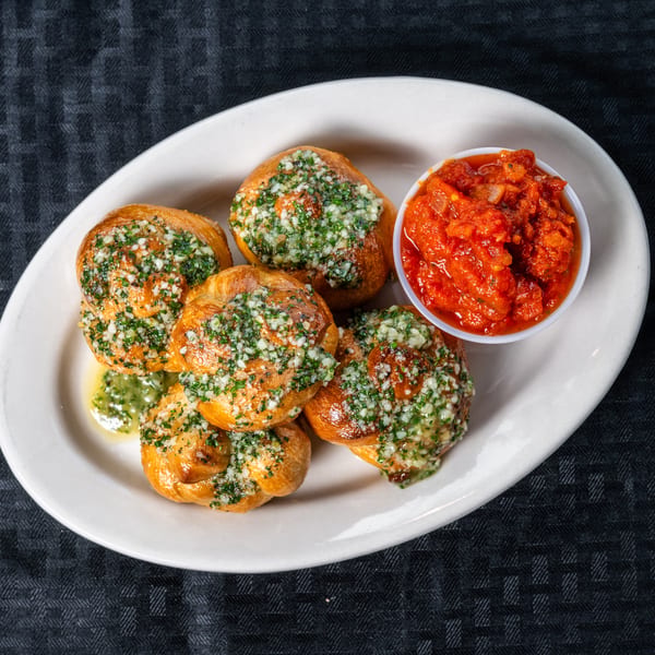 Six golden garlic knots topped with fresh herbs and garlic, served with a side of marinara sauce.