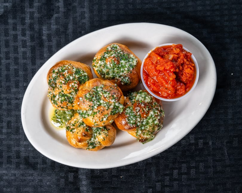 Six golden garlic knots topped with fresh herbs and garlic, served with a side of marinara sauce.