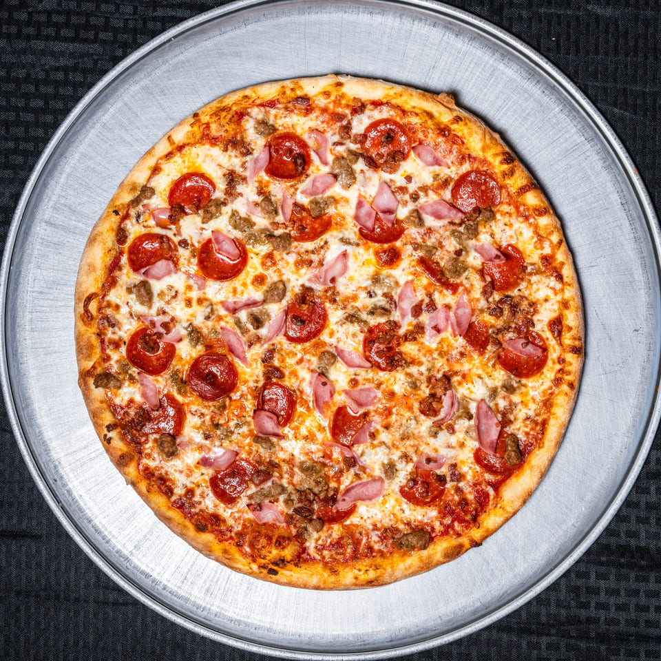 Round pizza topped with pepperoni, ham, and sausage on a silver metal pan.
